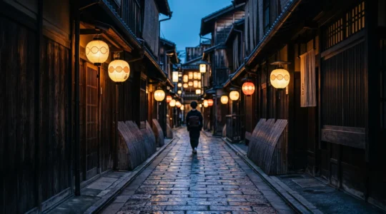 Ruelle pavee du quartier de Gion a Kyoto au crepuscule, lanternes allumees et silhouette discrete en kimono au loin